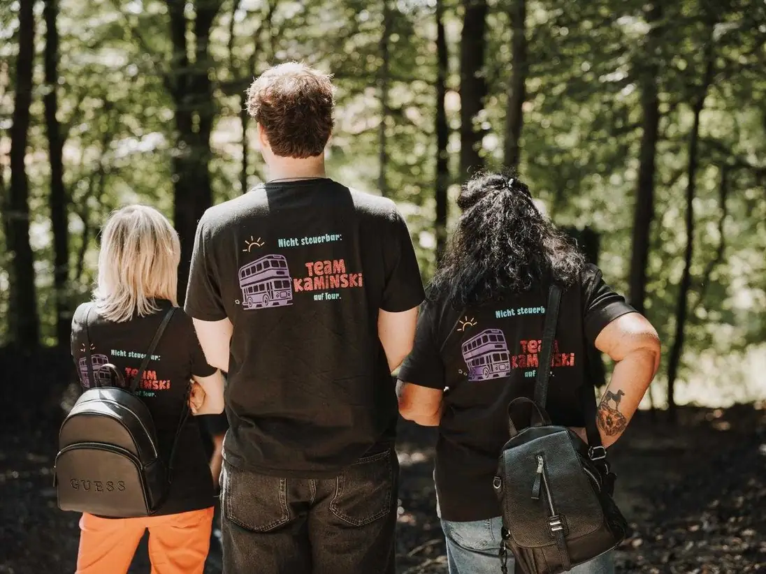 Drei Personen von hinten in schwarzen T-Shirts mit buntem Rückendruck, ein Doppeldeckerbus und ein Schriftzug